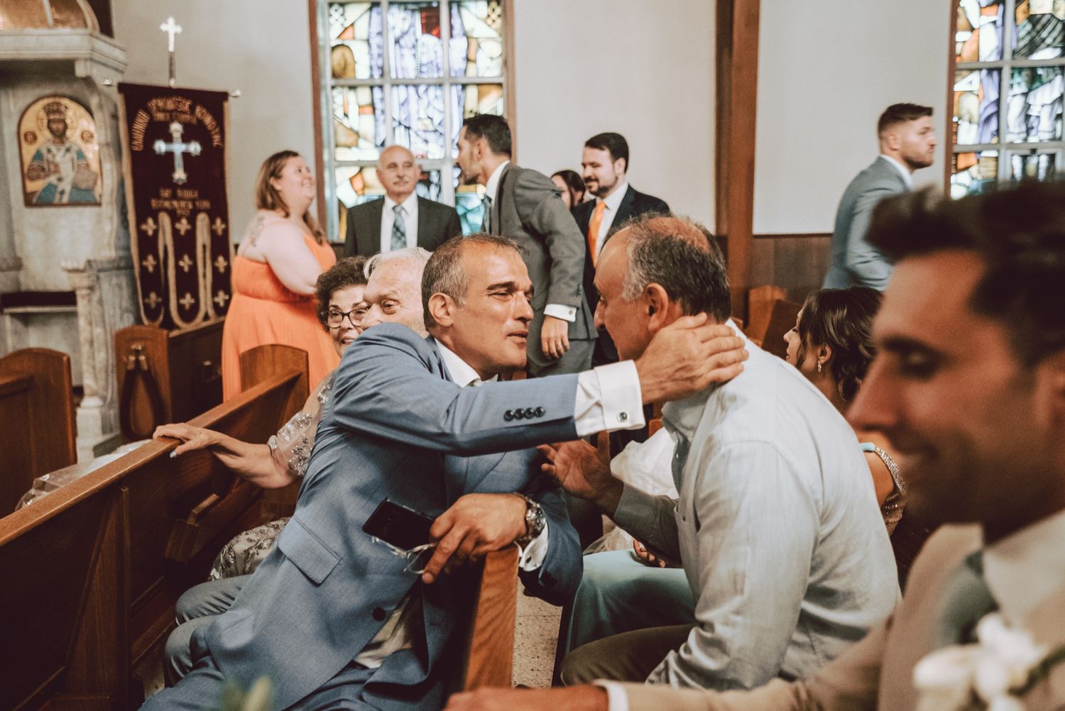 Men in suits greeting each other warmly in church pews during a wedding ceremony with other guests seated nearby.