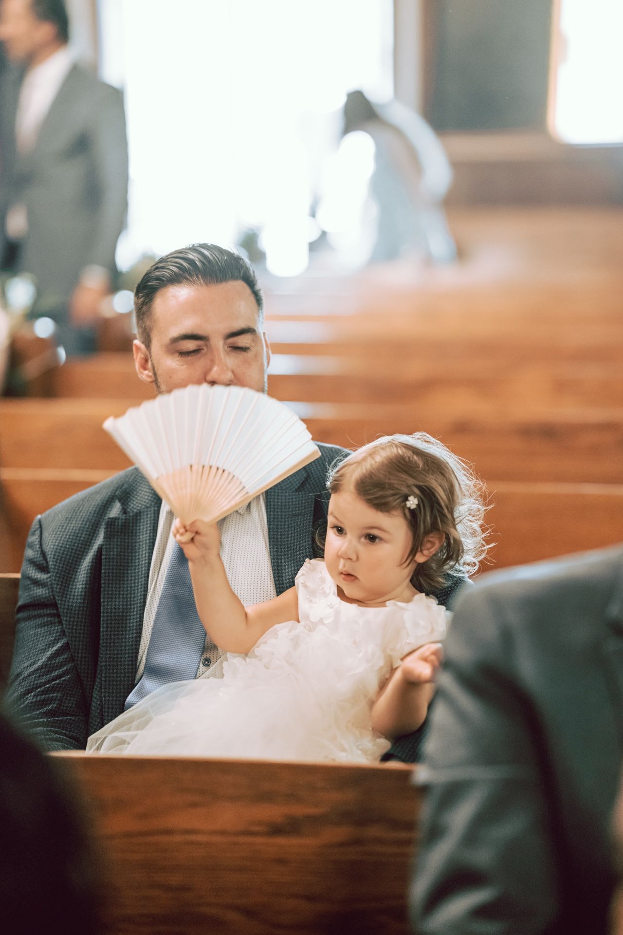 A man in a suit holds a white hand fan with a little girl in a white dress sitting on his lap in a church pew.