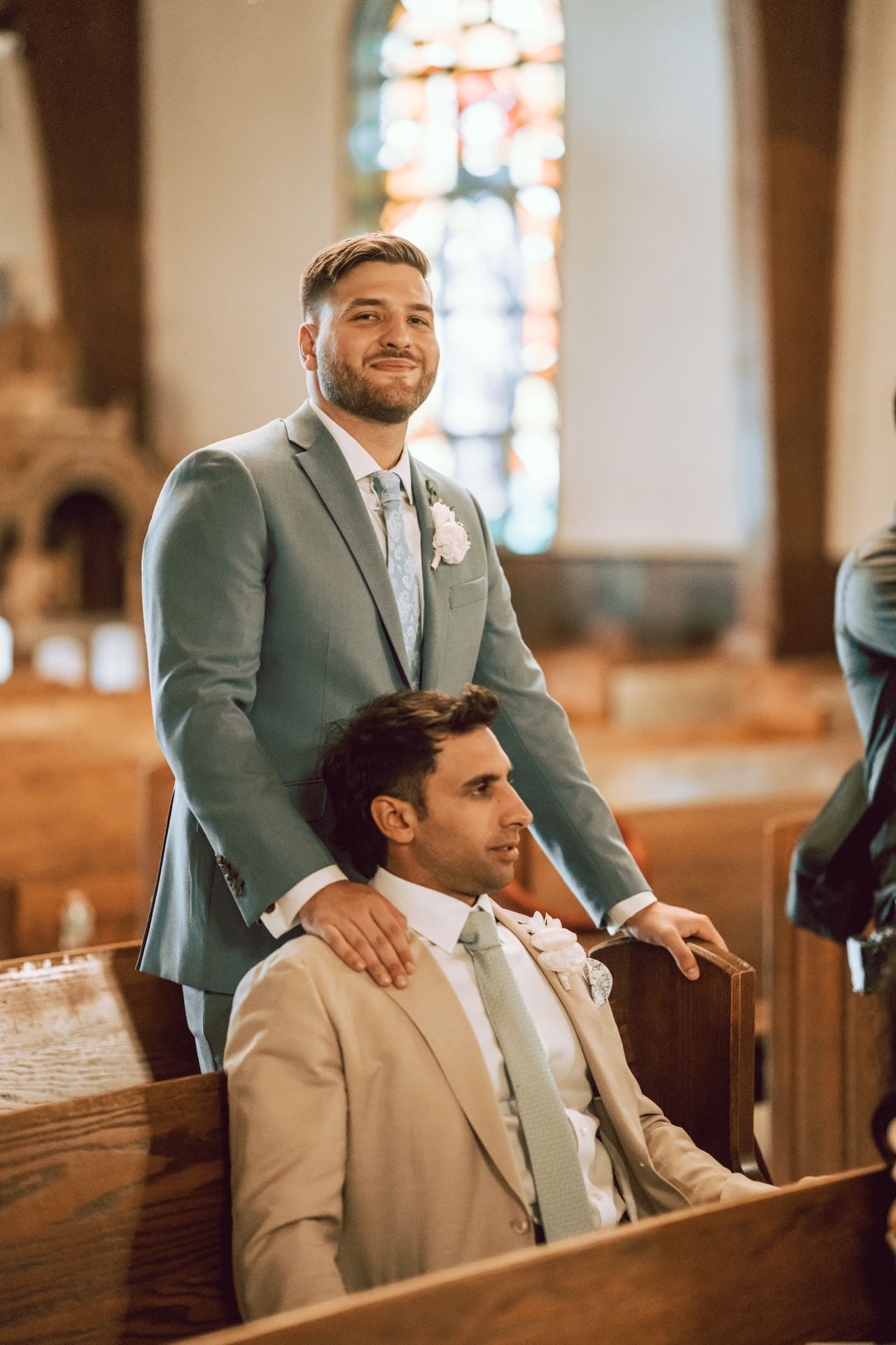 Two groomsmen in gray and beige suits sitting in church pews during a wedding ceremony with stained glass windows.