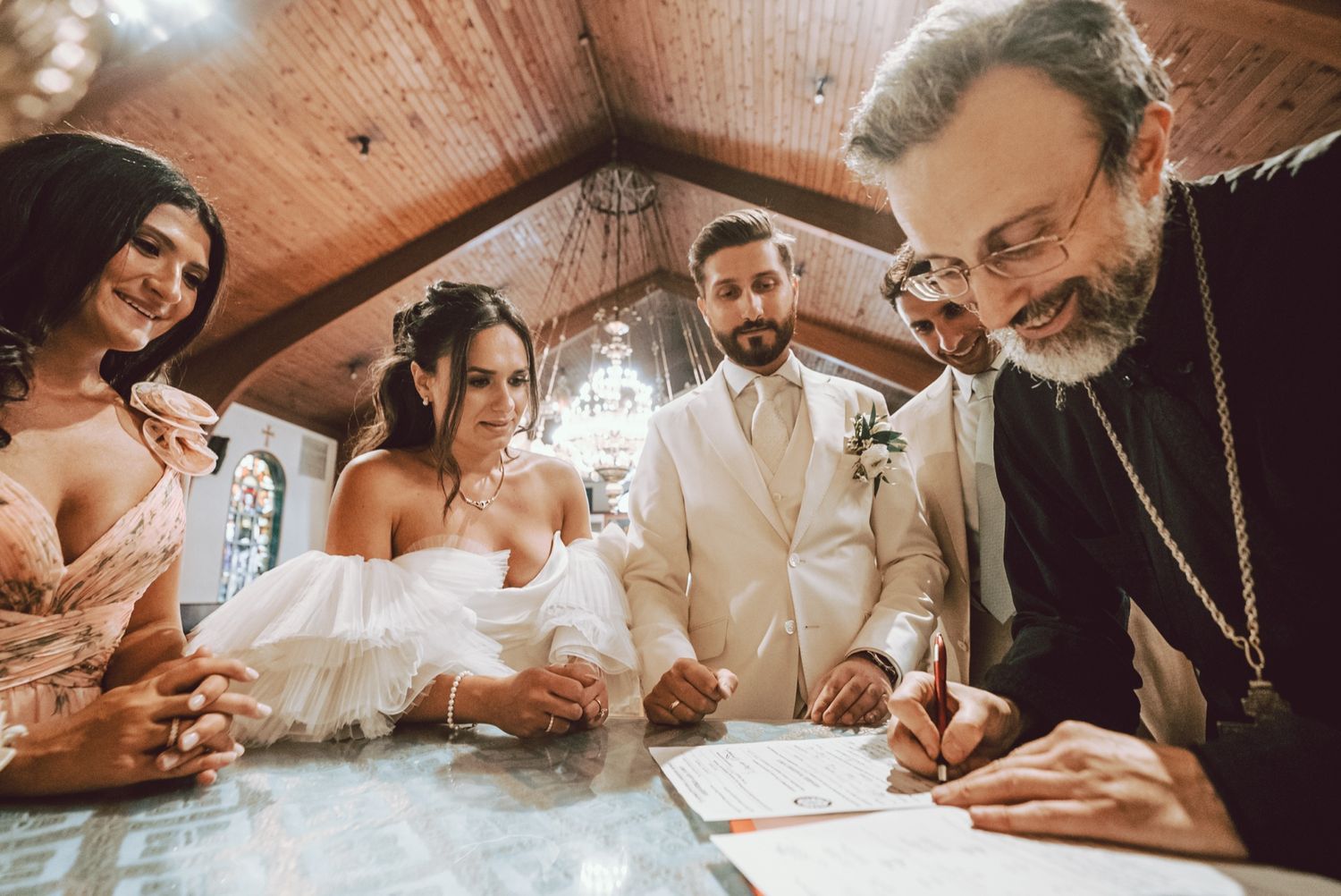 Bride and groom sign wedding documents with a bearded officiant and guests watching inside a wooden-ceiling chapel.