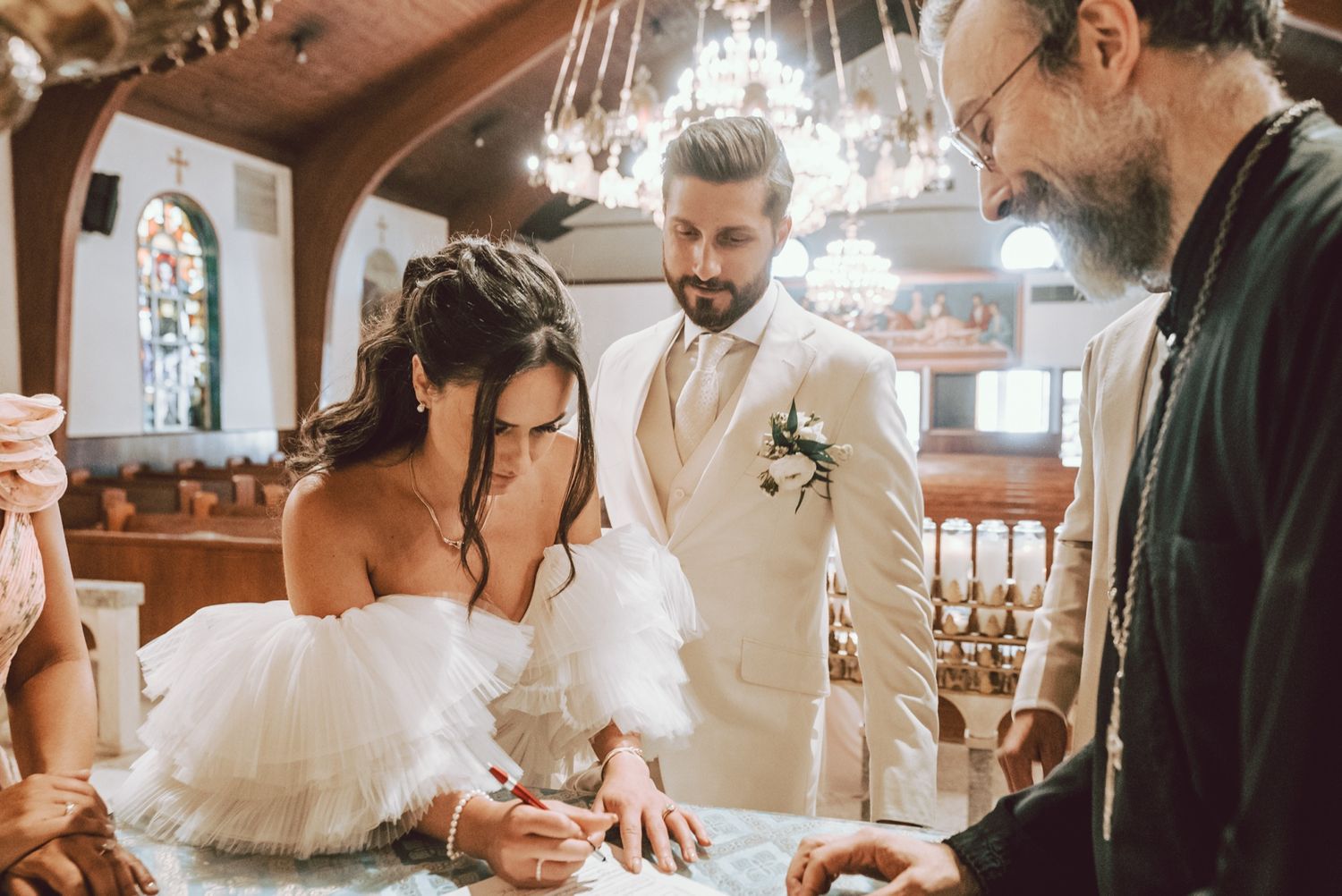 Bride in off-shoulder white dress signs wedding registry with groom in ivory suit and officiant in a rustic church.