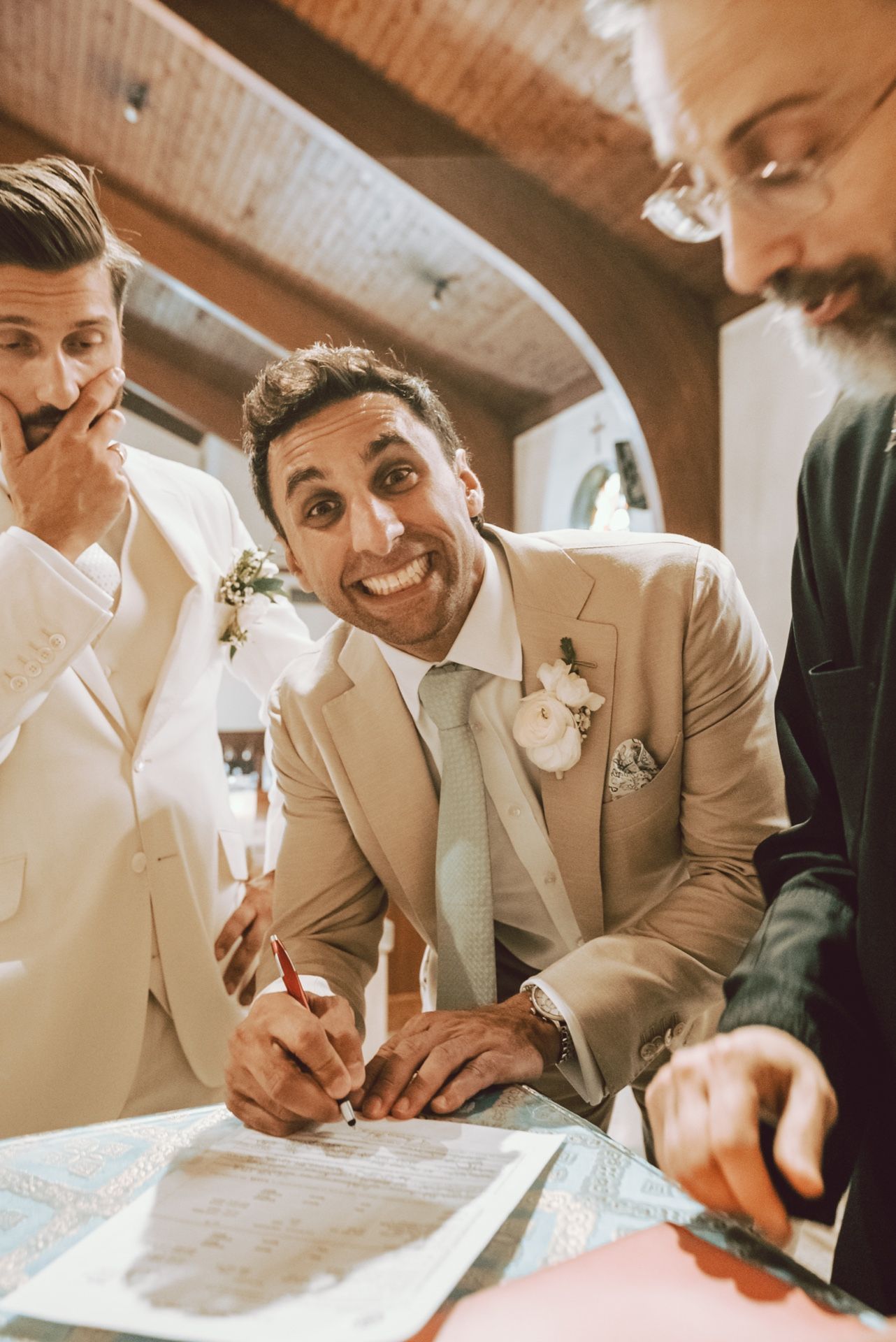 Excited groom in beige suit signing marriage certificate with groomsmen at wedding ceremony.