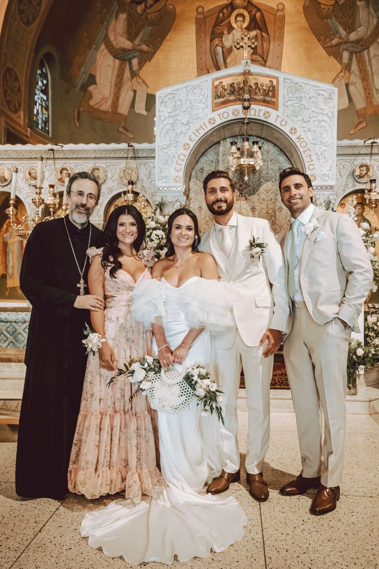 Bride and groom pose with guests and Orthodox priest inside a beautifully decorated church on their wedding day.
