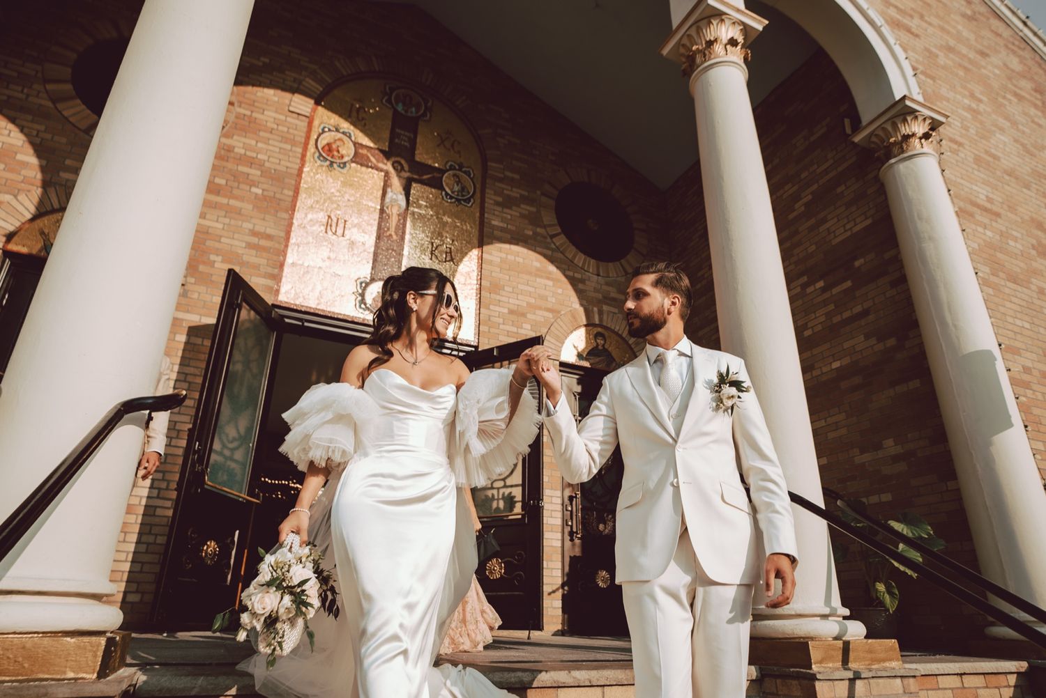 Bride and groom walk down church steps, she in a white gown with feathers, he in a white suit, holding hands.