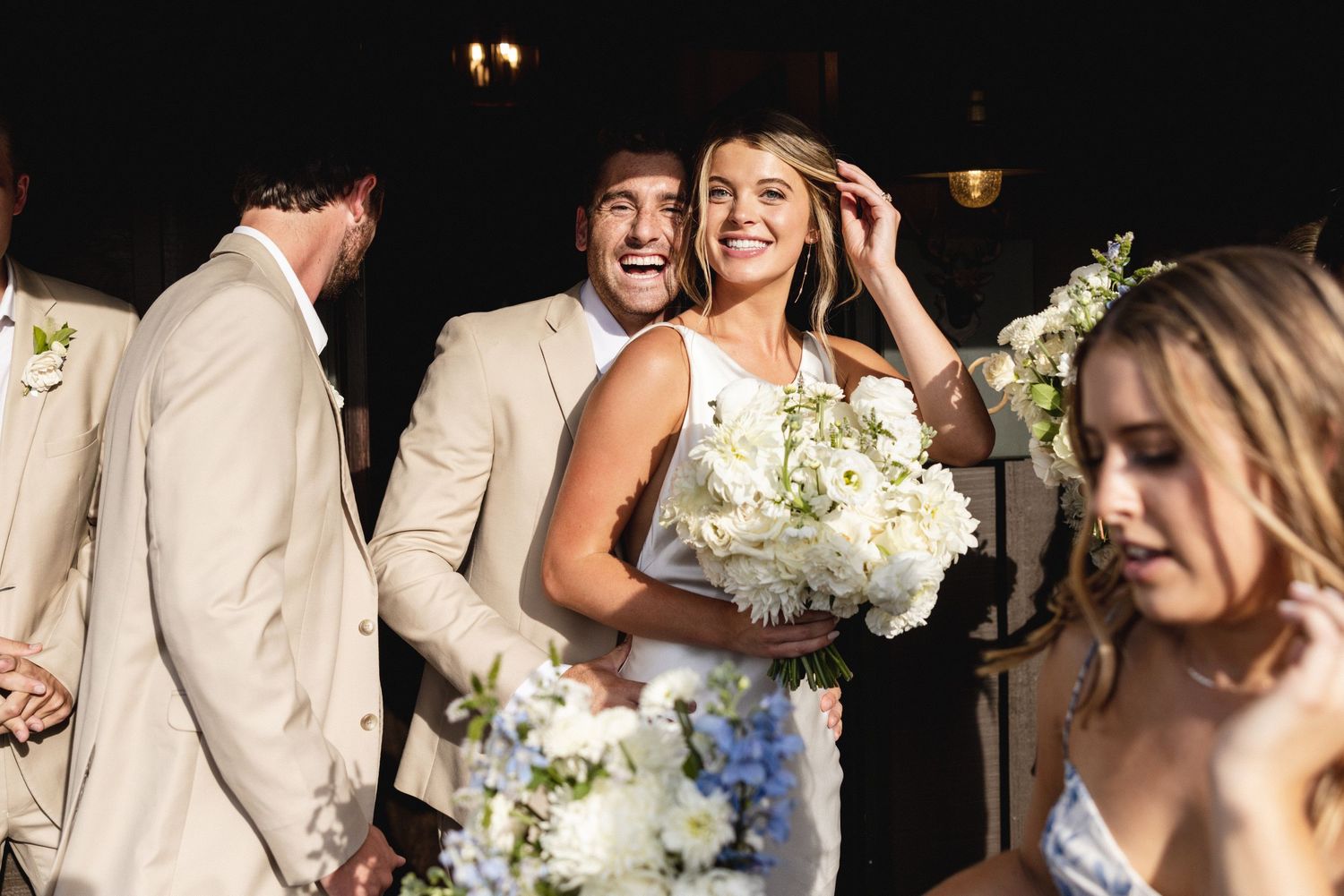 Joyful bride holding white and blue bouquet laughs with groom in tan suit during outdoor wedding celebration.