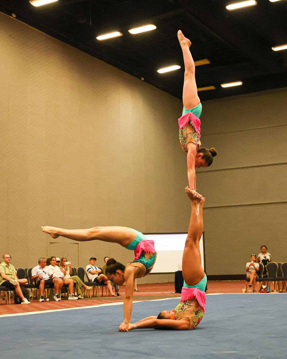 Three gymnasts in colorful costumes perform an acrobatic balancing act during a competition in an indoor arena.