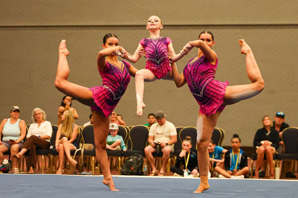 Three gymnasts in pink leotards perform synchronized leg holds at a gymnastics competition indoors.