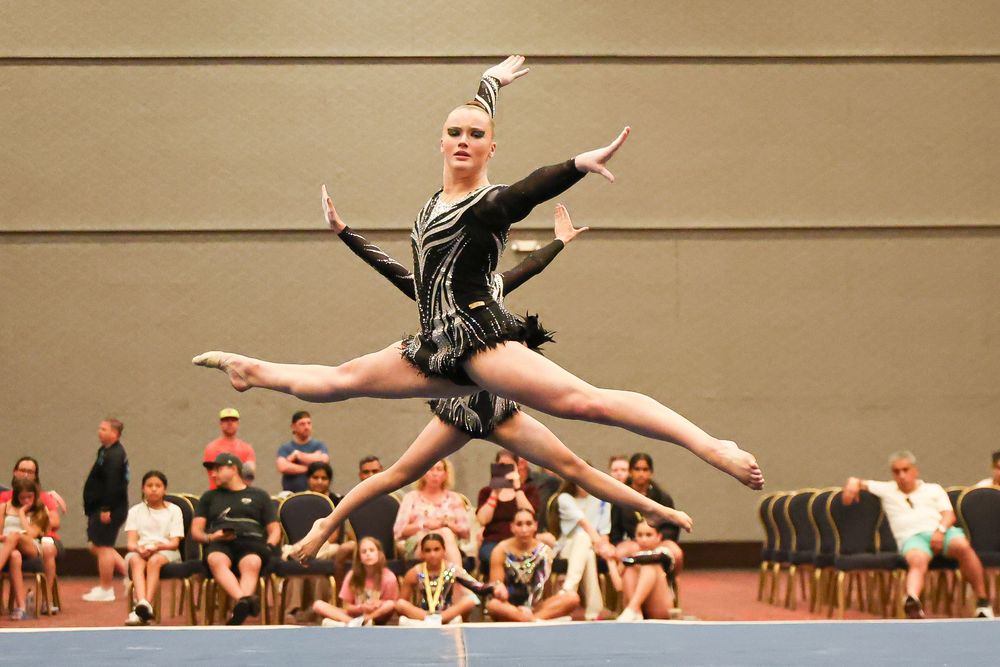 Gymnast performing a split leap in black costume at indoor competition with spectators watching.
