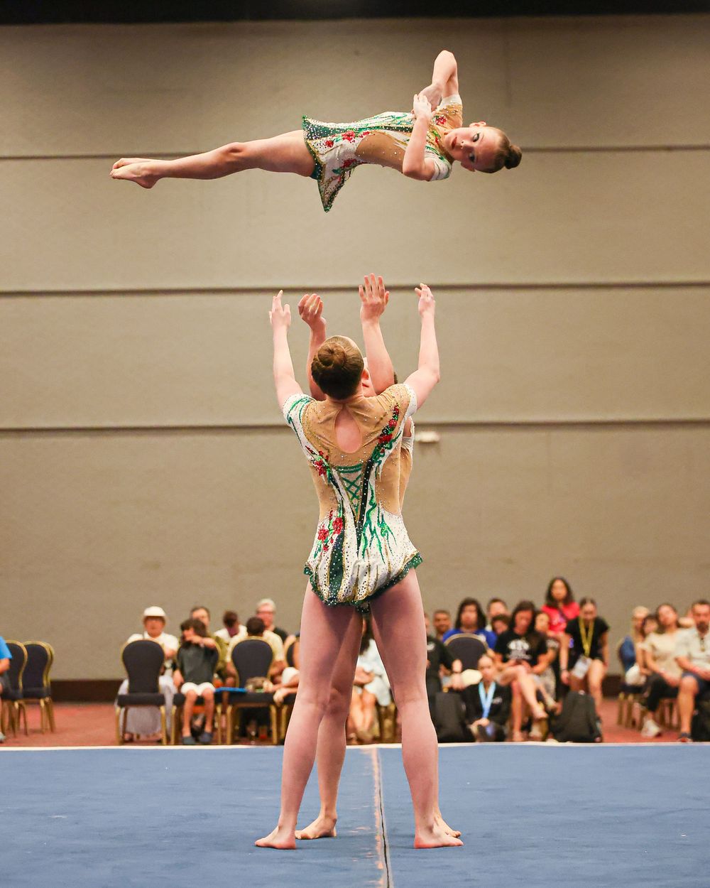 Two acrobatic gymnasts perform a balance stunt, one lifted horizontally above the other in matching floral leotards.