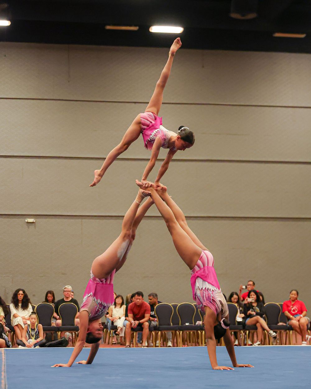 Three gymnasts in pink leotards perform an acrobatic balance pyramid at an indoor gymnastics competition.