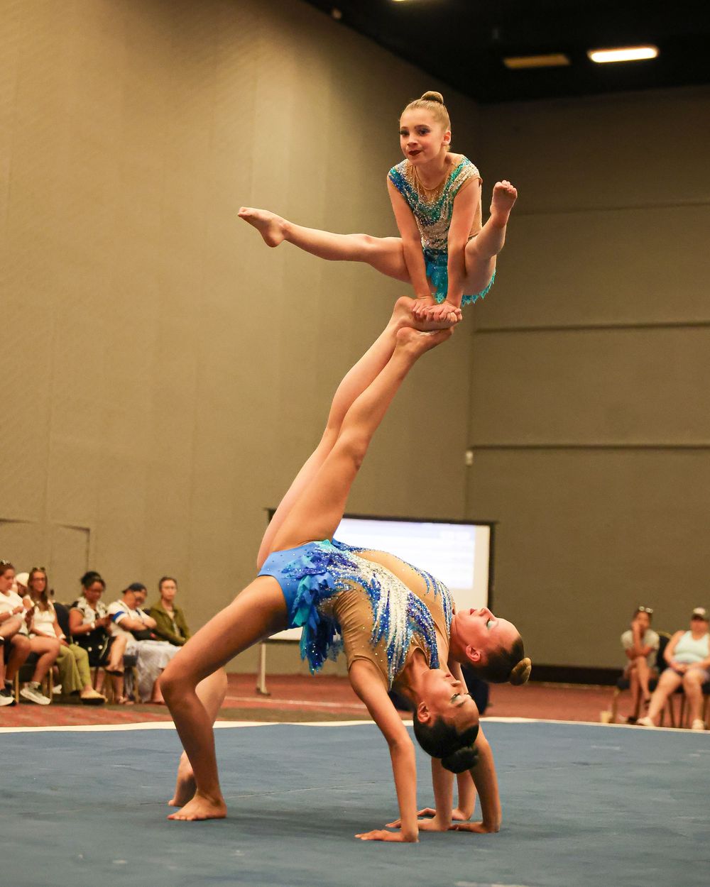 Three acrobatic gymnasts performing a human pyramid stunt in blue sequined leotards on a blue competition mat.