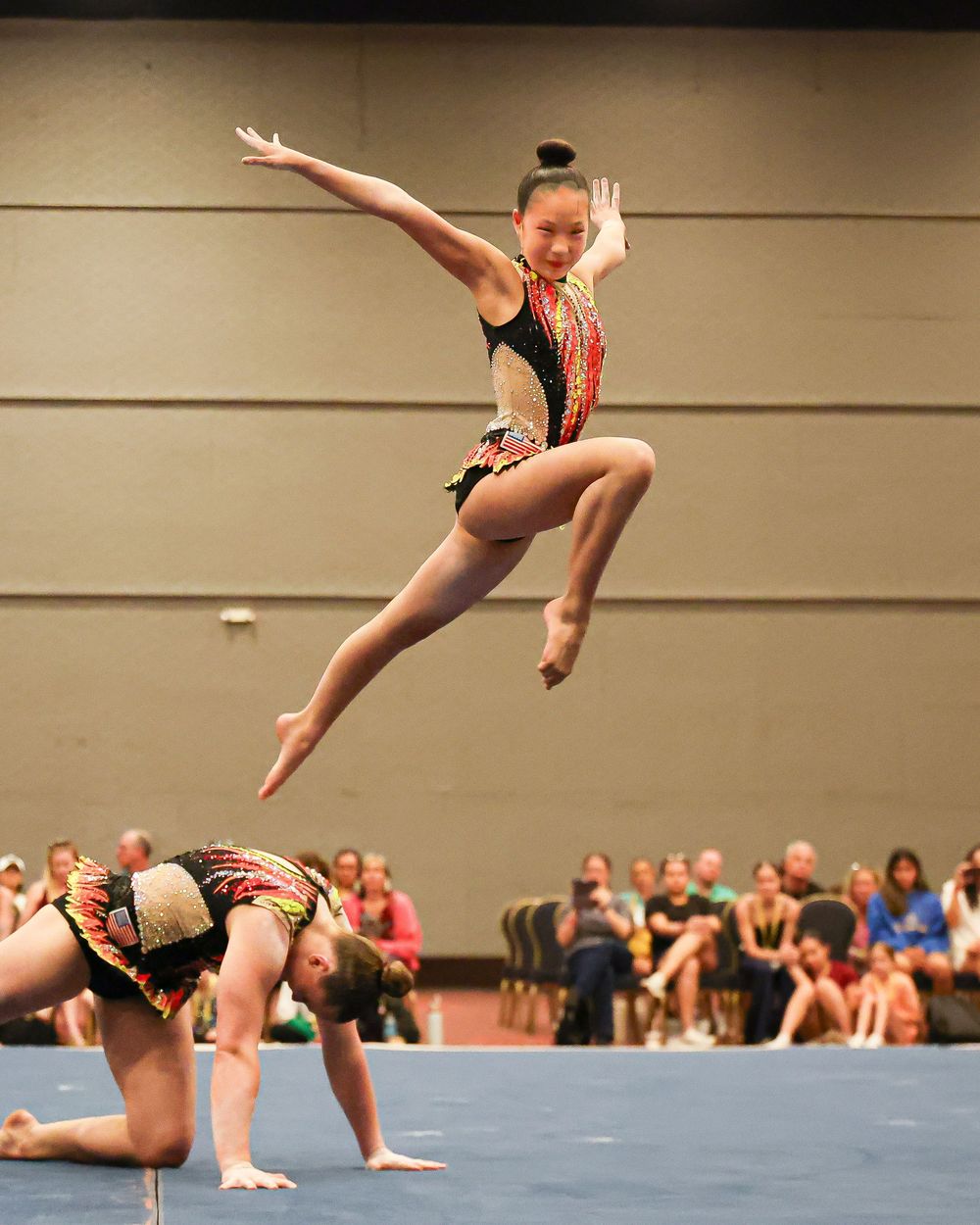 Young gymnast leaps mid-air in colorful leotard during acrobatic floor routine at indoor gymnastics competition.