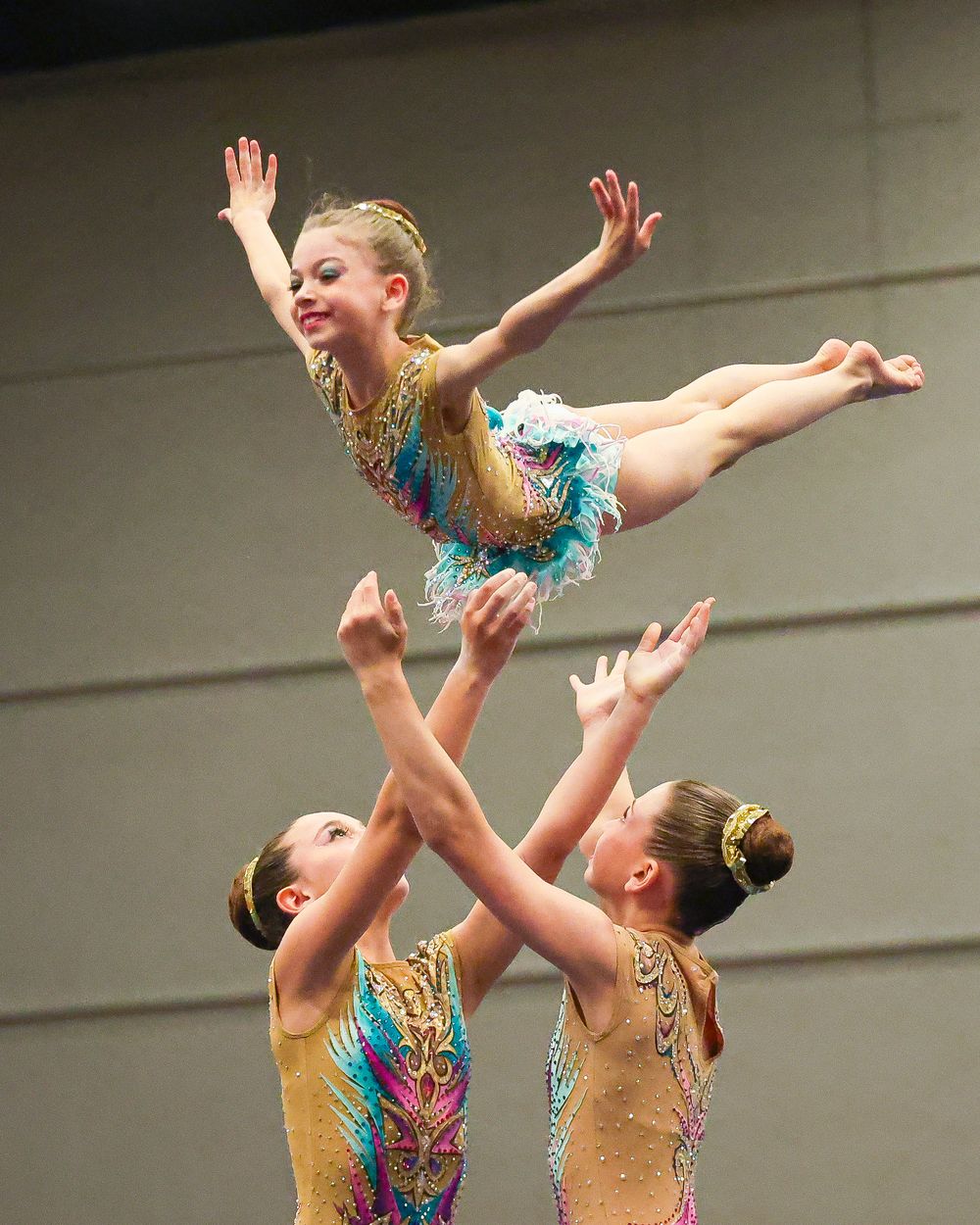 Three young gymnasts perform acrobatic routine, with one girl lifted overhead in a graceful pose wearing colorful leotards.