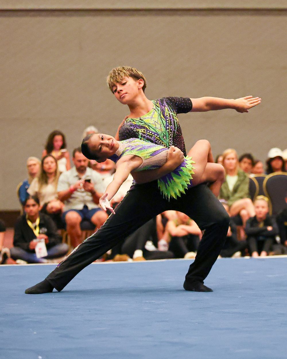 Two acrobatic gymnasts perform a dynamic lift routine in colorful costumes at a competitive gymnastics event.