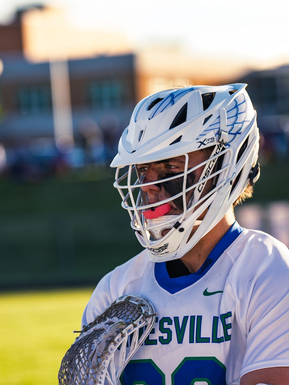 Janesville lacrosse player in a white jersey and helmet with black eye paint and a pink mouthguard, stick in hand at golden hour.