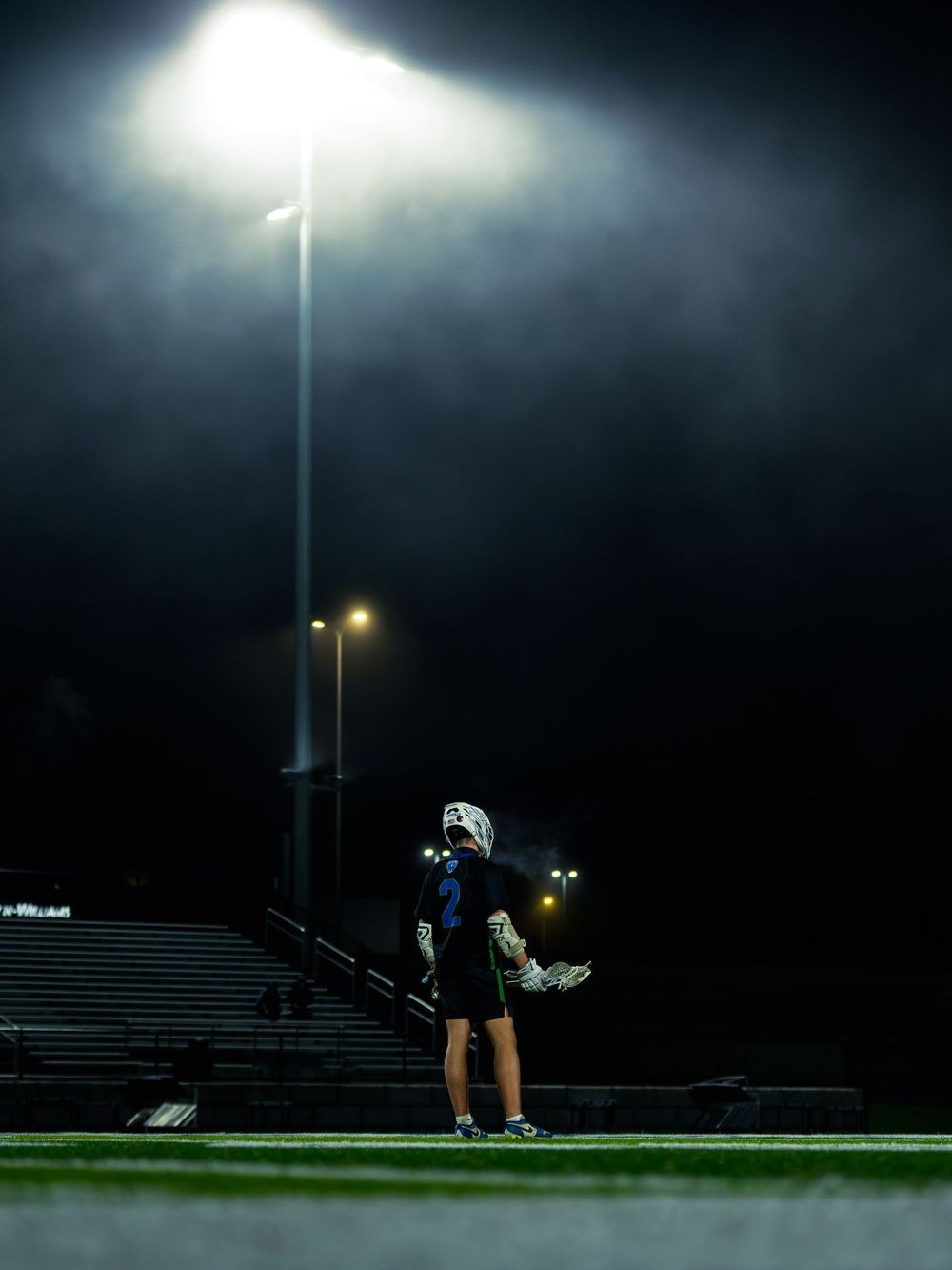 Janesville lacrosse player number 2 stands alone on the field under a glowing stadium light on a misty night, breath visible in the cold.