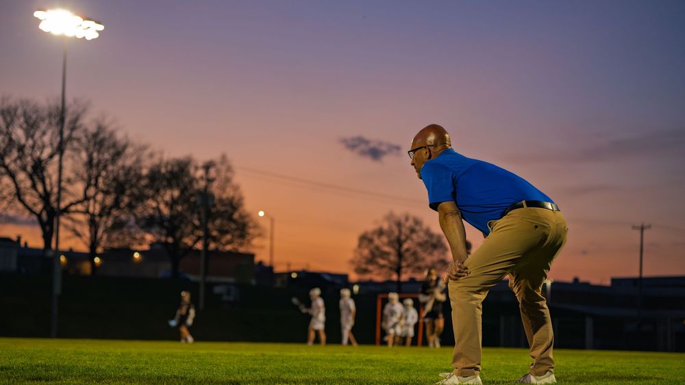 A Janesville lacrosse assistant coach leans on his knees to watch the game from the sideline at sunset as players warm up behind him.