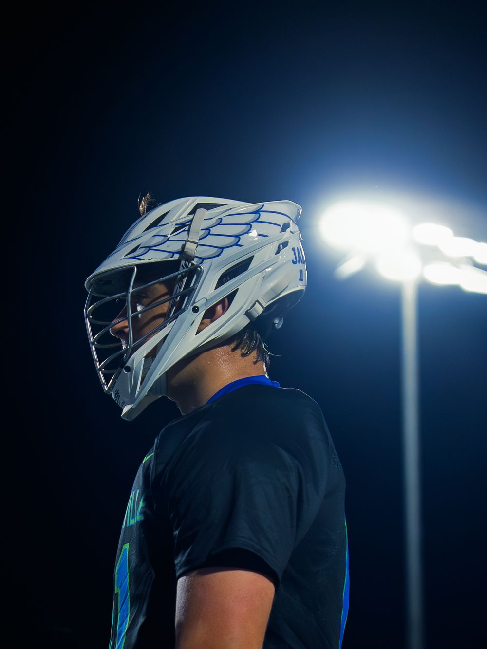 Janesville lacrosse player in profile wearing a white helmet and black jersey, lit against the dark by a bright stadium light at night.