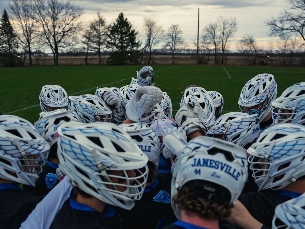 The Janesville lacrosse team huddles close in white helmets, raising gloved hands together before the game under a gray sky.