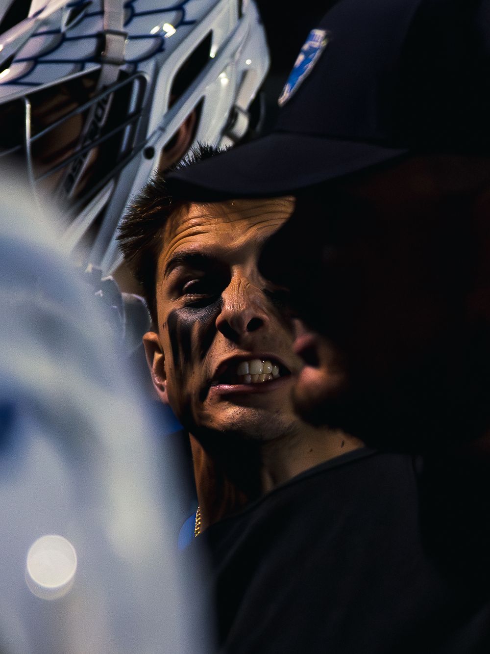 Intense close-up of a Janesville lacrosse player in a cap with black eye paint, teeth gritted, a helmet held close beside his face.