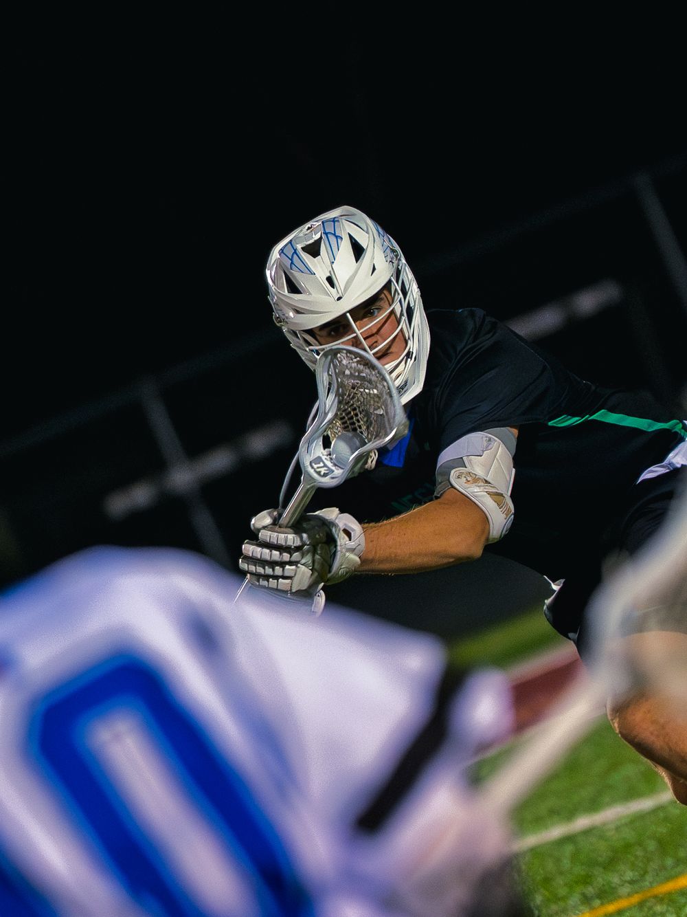 Janesville lacrosse player in black crouches over his stick at a faceoff, eyes locked on the ball against the opponent at night.