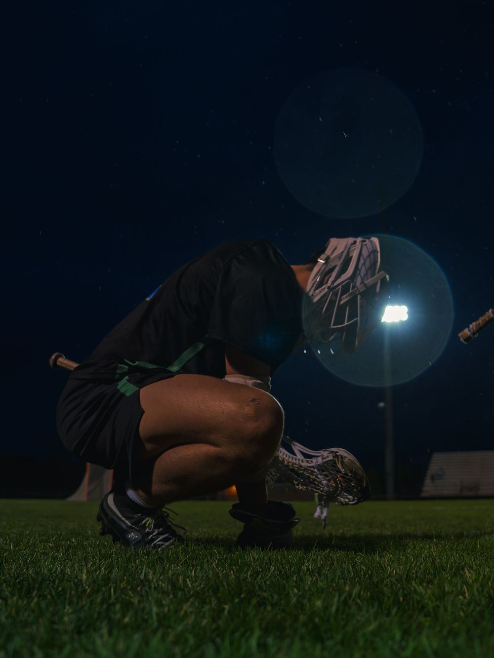Janesville lacrosse player crouches low on the grass with stick in hand, backlit by a stadium light and lens flare at night.