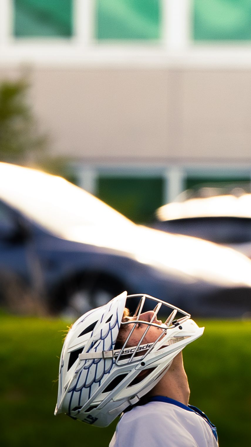 Janesville lacrosse player in a white helmet tilts his head back and looks skyward, lit by warm light with a blurred building behind.