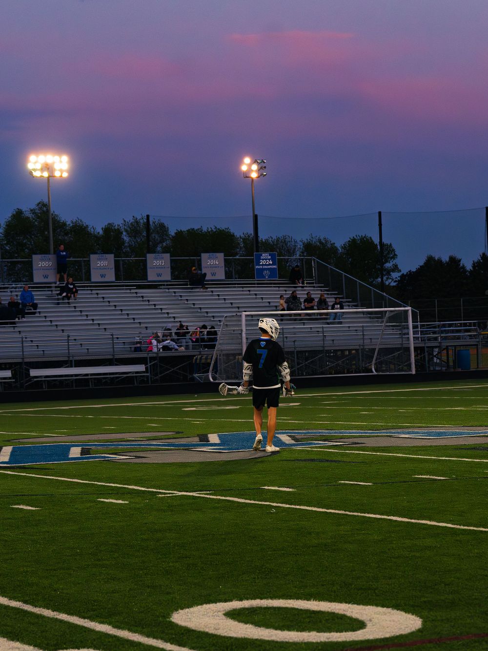 Janesville lacrosse player number 7 walks alone across the field toward the bleachers under stadium lights at a pink and purple dusk.
