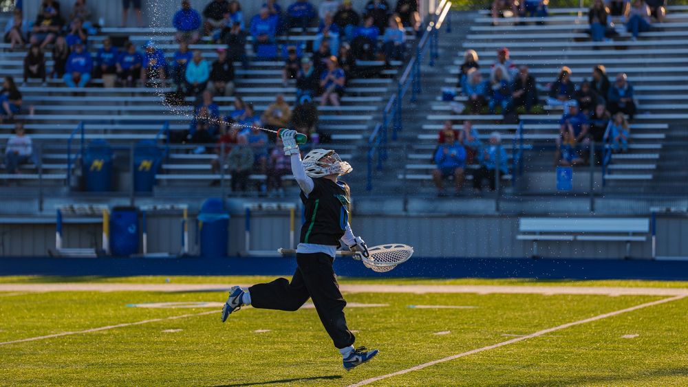 Janesville lacrosse goalie sprints down the field spraying a water bottle overhead in celebration as fans watch from the bleachers.