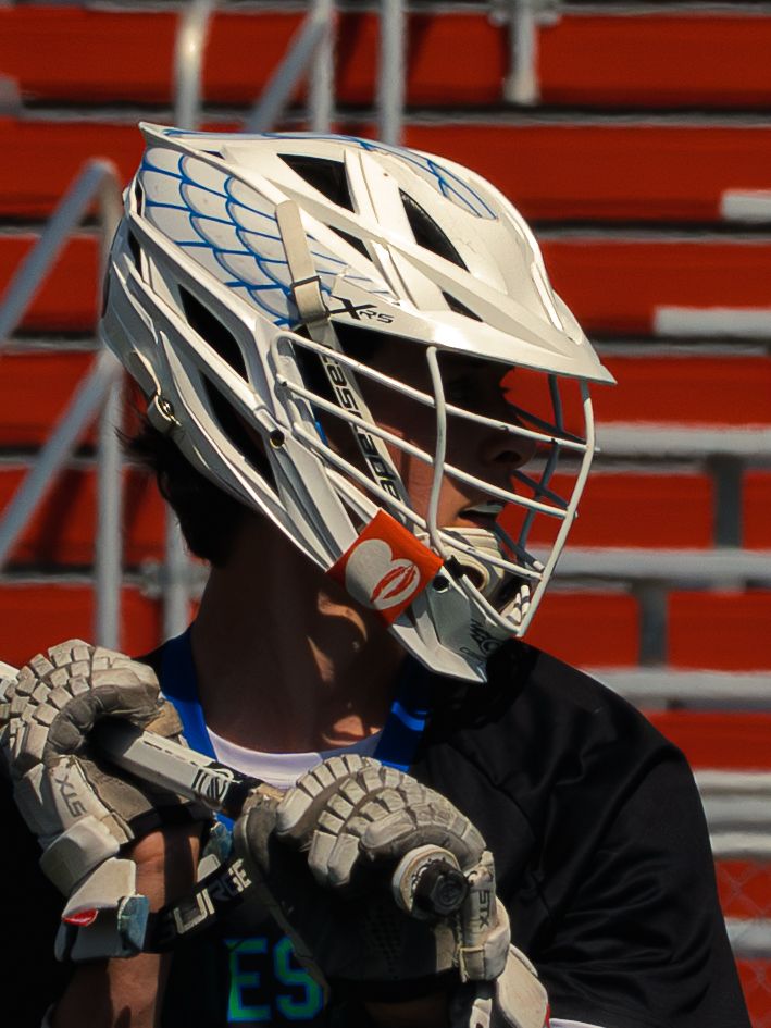 Side profile of a Janesville lacrosse player in a white helmet and black jersey, gloved hands on his stick, red bleachers behind.
