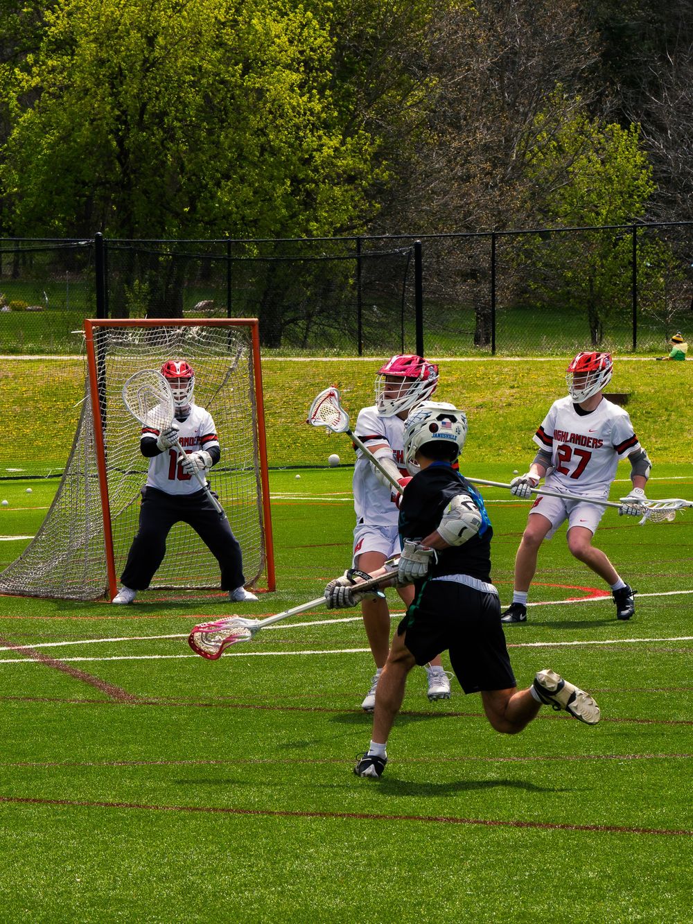 Janesville lacrosse player in black drives toward the net as the Highlanders goalie and defenders set to block in bright daylight.