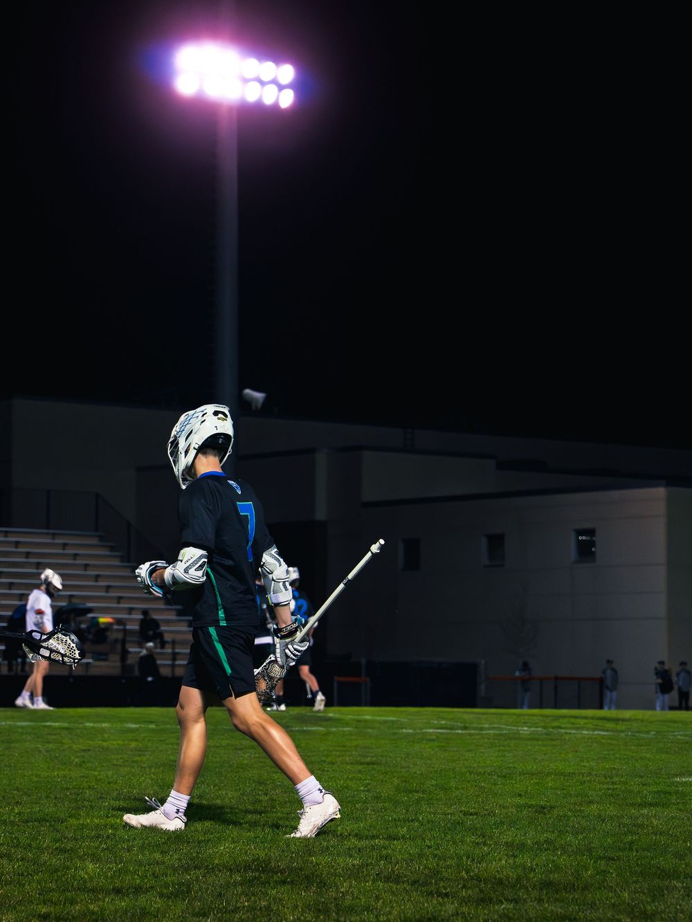 Janesville lacrosse player number 7 walks across the field with a long defensive stick under a bright stadium light at night.