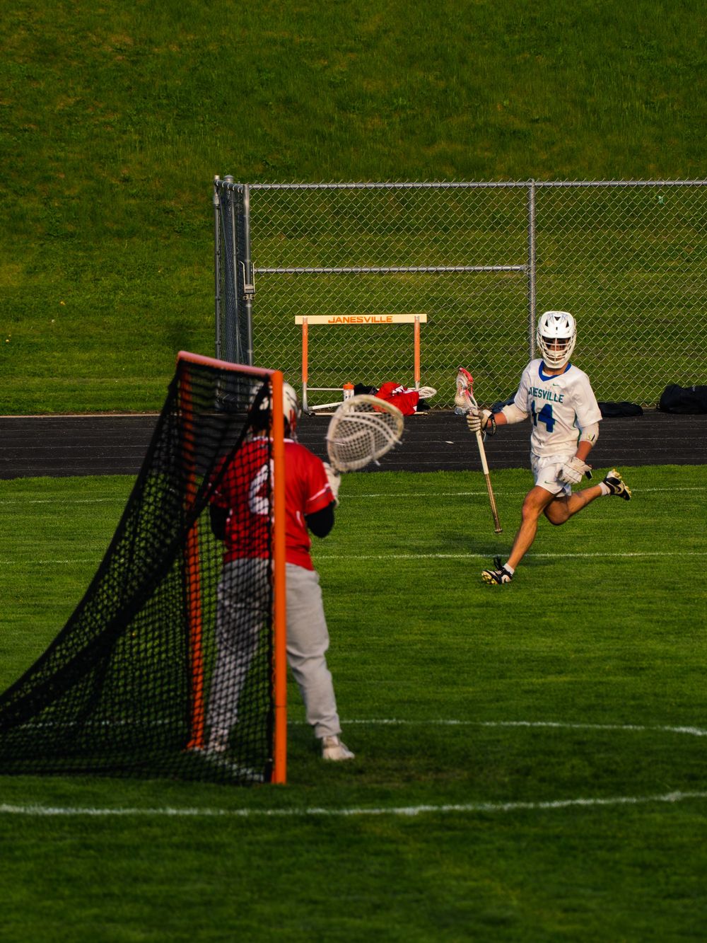 Janesville lacrosse player number 14 in white races toward the net with the ball as a red-jersey goalie sets to defend in evening light.