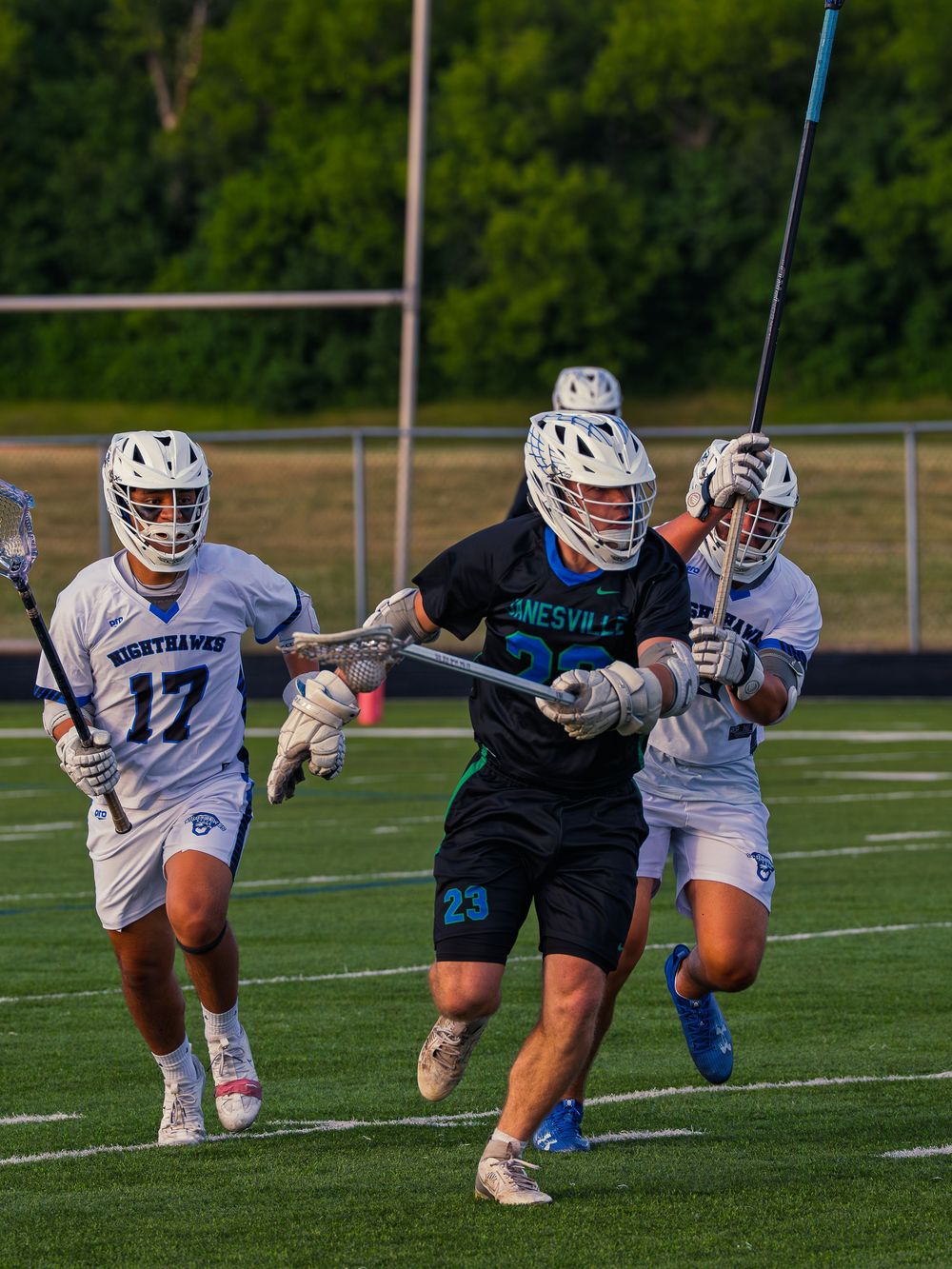 Janesville lacrosse player number 23 in black drives the ball upfield between two white-jersey Nighthawks defenders in evening light.