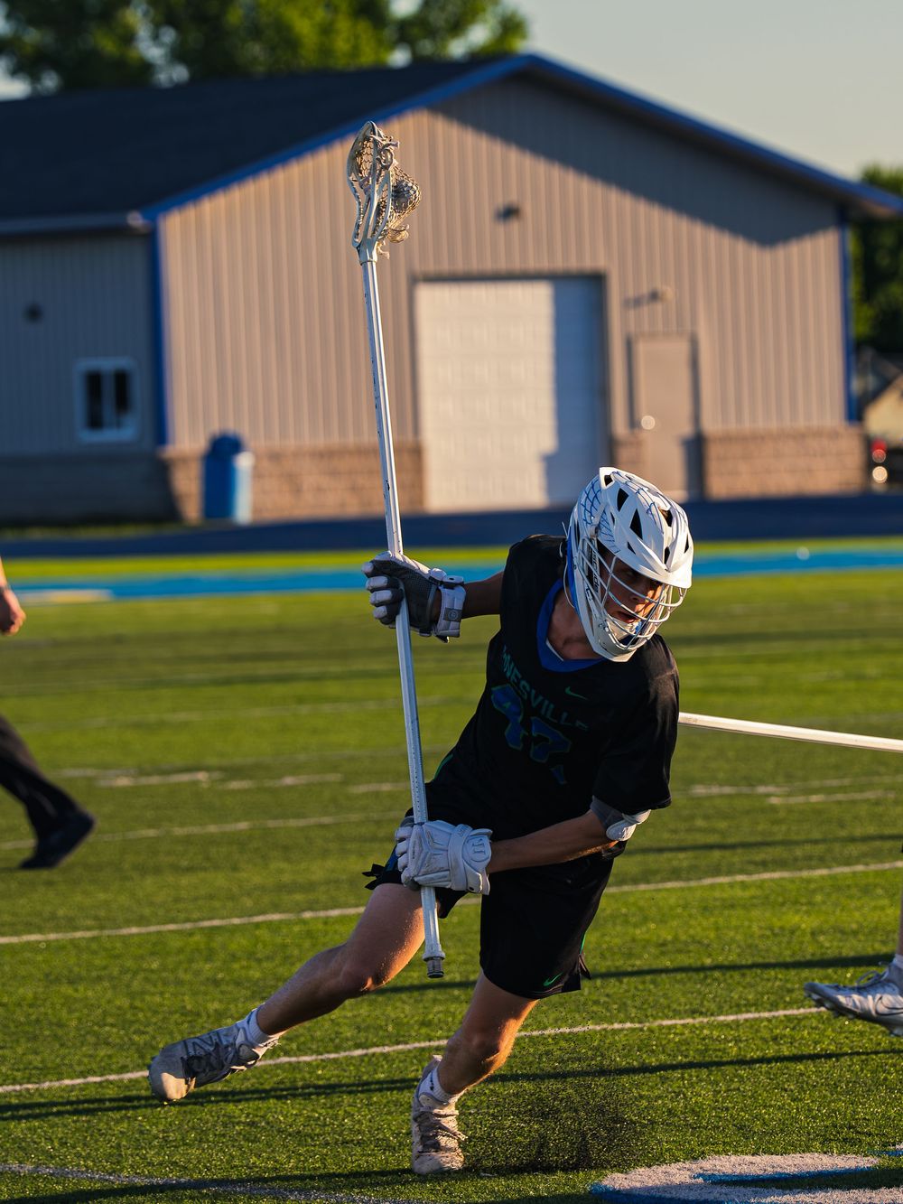 Janesville lacrosse player in black drives upfield with stick held high in golden evening light, a metal building behind him.