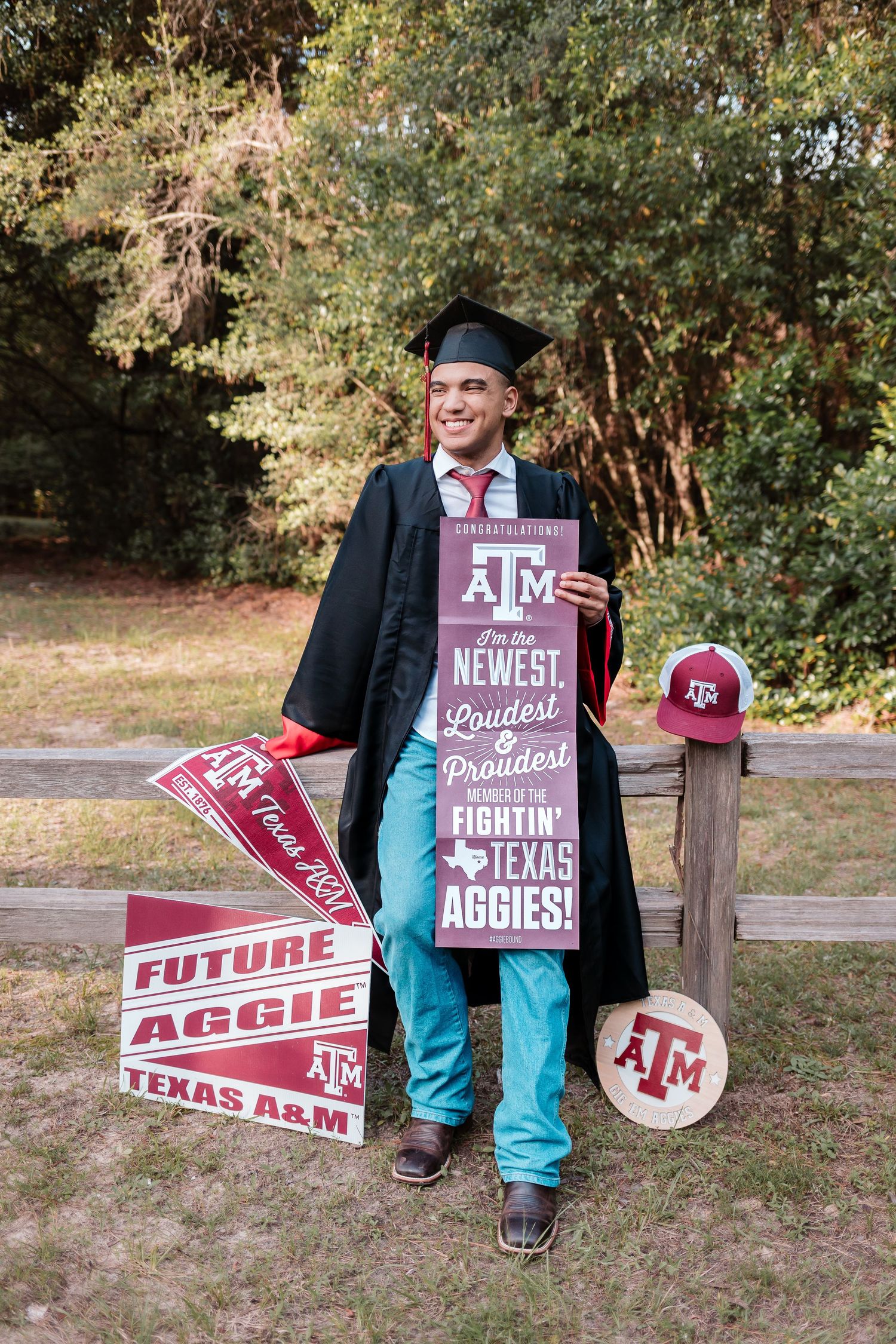 Texas A&M graduate smiles in cap and gown holding Aggies signs and pennants outdoors.