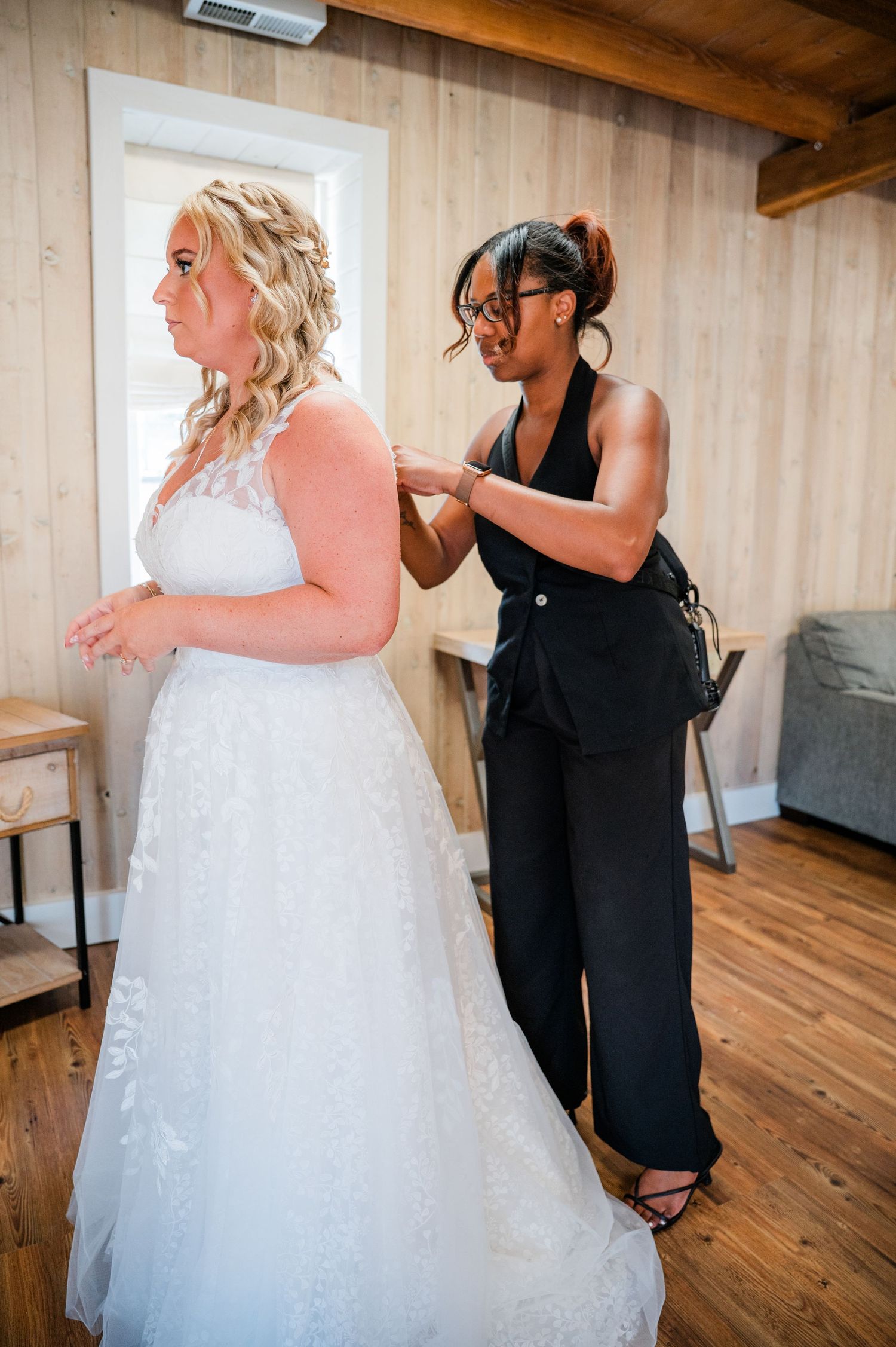 A wedding planner from Cherished Events and Designs helps a bride in a white lace gown get dressed in a rustic wooden room.