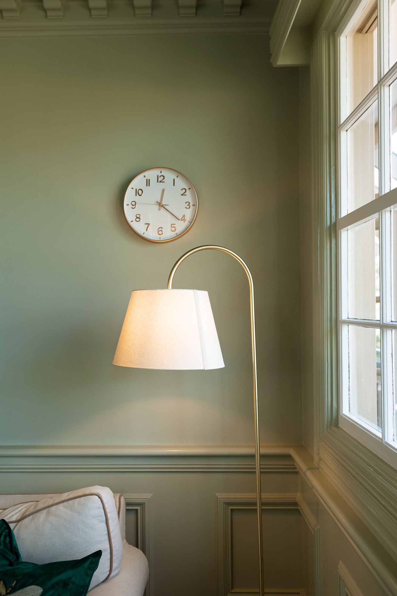 Elegant living room corner with sage green walls, arc floor lamp, round wall clock, and white sofa near a bright window.