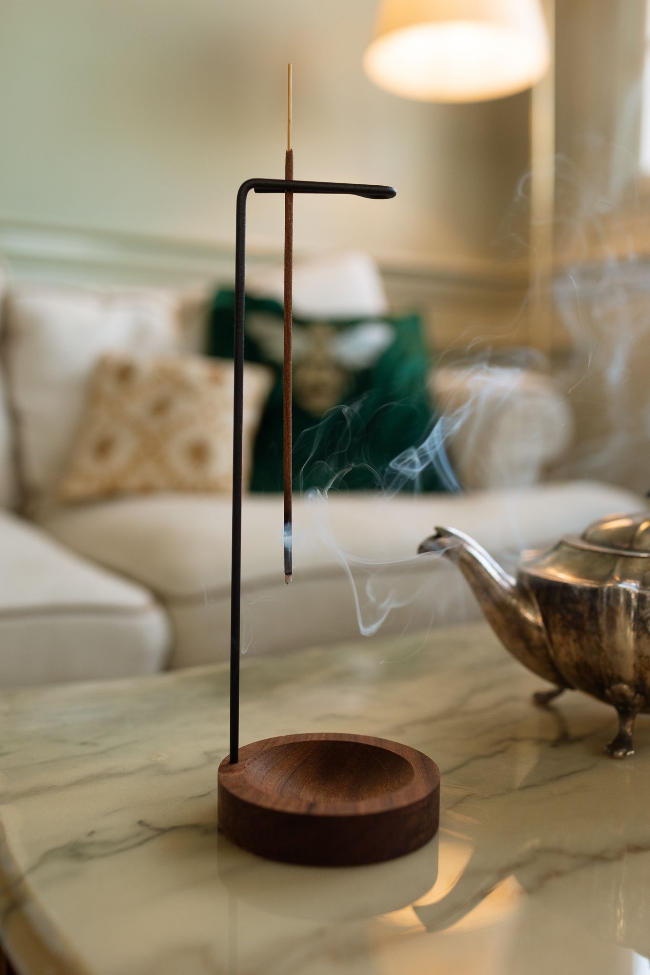 Minimalist incense holder with burning stick on marble table, smoke curling upward, beside silver teapot in cozy living room.