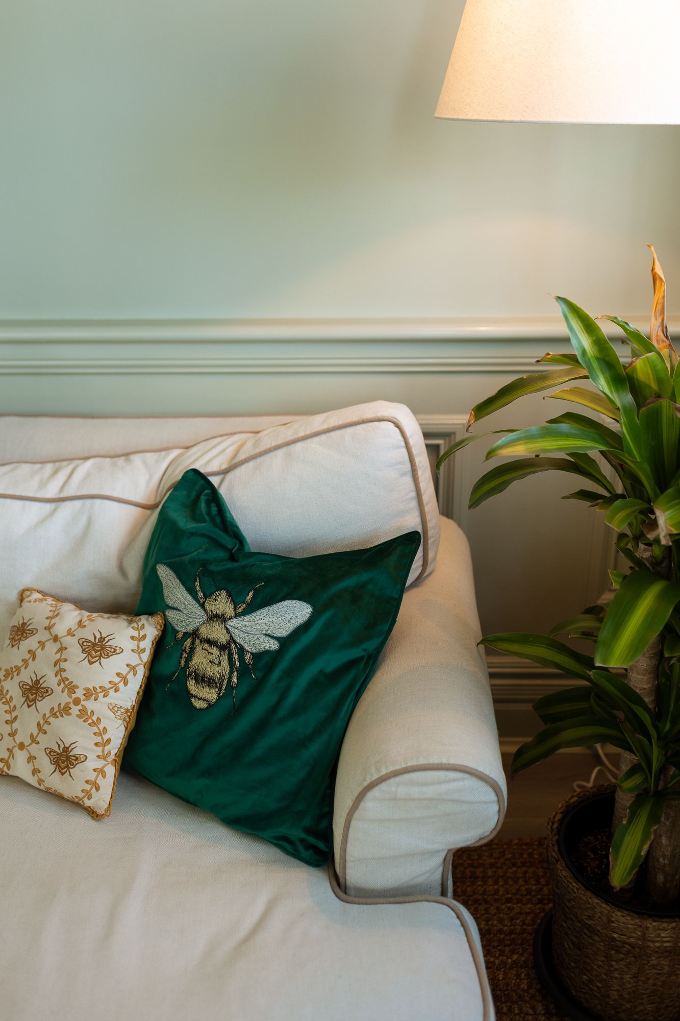 White sofa with green velvet bee embroidered cushion and floral pillow, beside a houseplant and lamp.