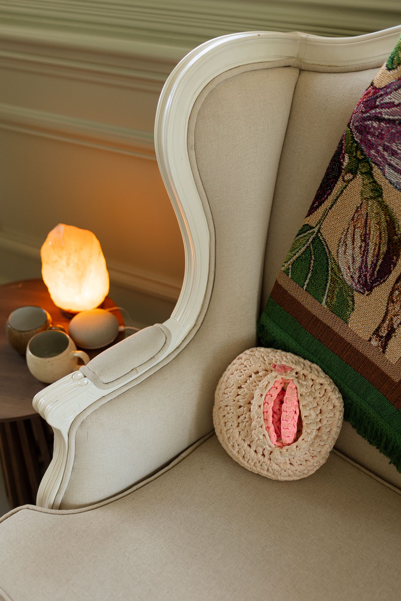 Cream wingback chair with crochet vulva pillow, floral tapestry cushion, and glowing salt lamp on side table.