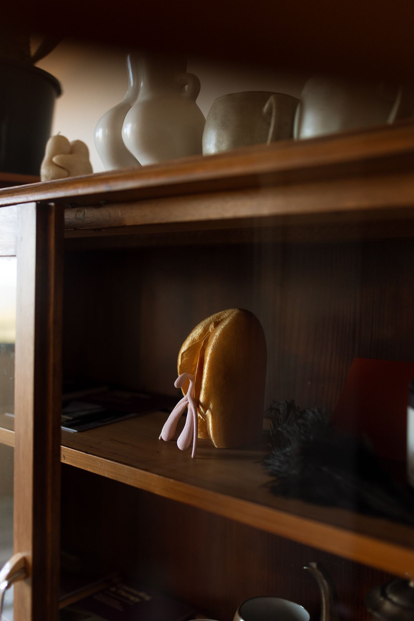 Wooden display cabinet with white ceramic vases on top shelf and a golden decorative object with pink ribbon on middle shelf.