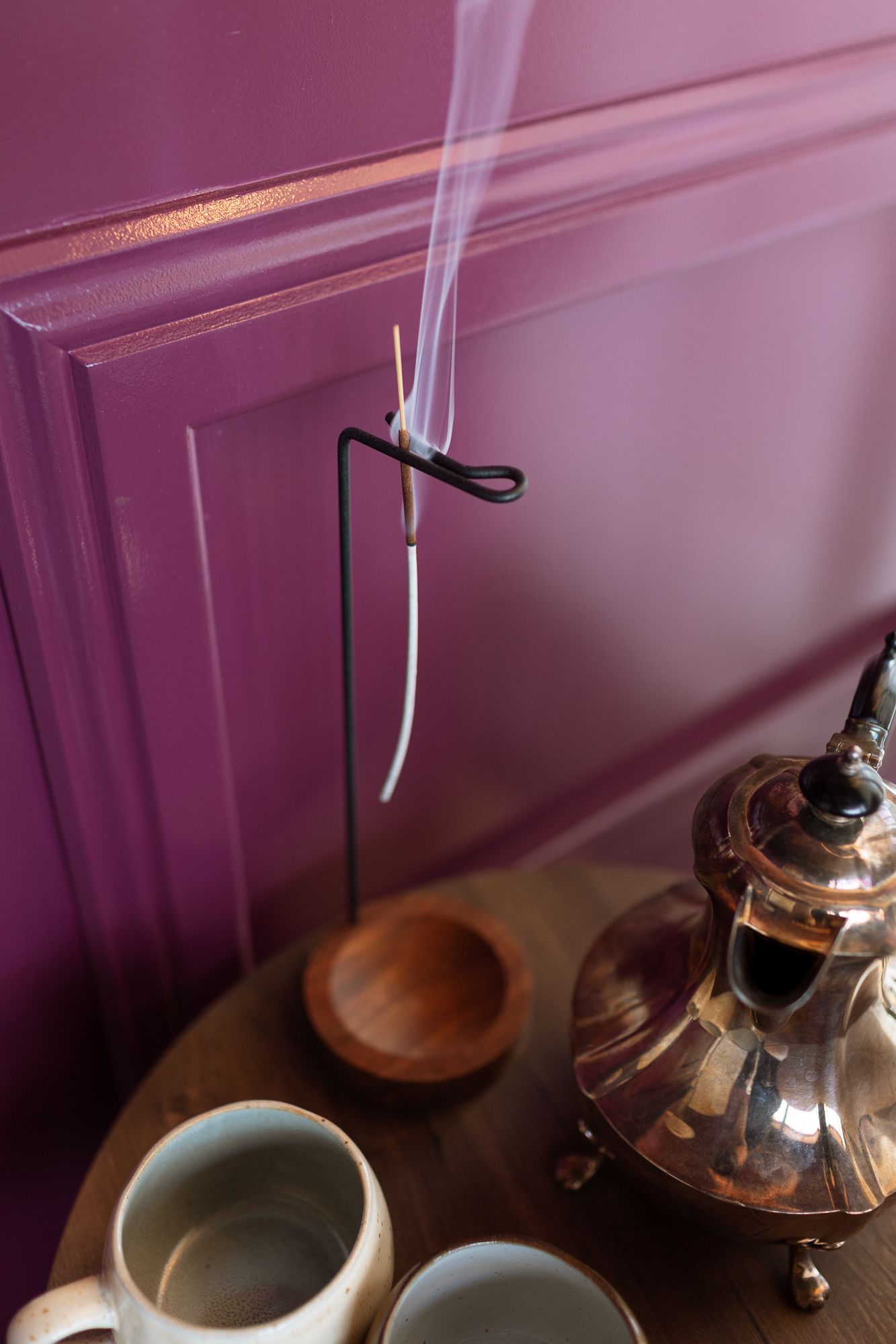 Burning incense stick on black holder with smoke rising, copper teapot and ceramic mugs on wooden tray against purple wall.