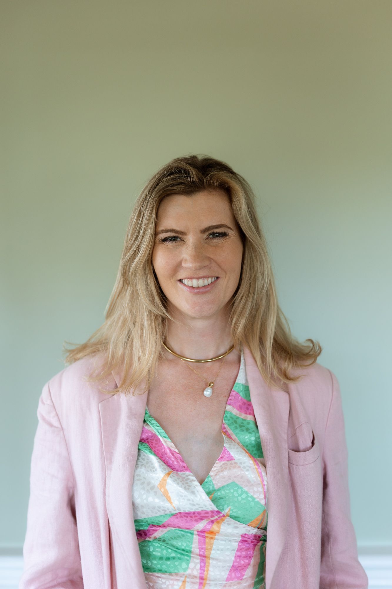 Smiling blonde woman in pink blazer and colorful dress wearing gold necklace against mint green wall.