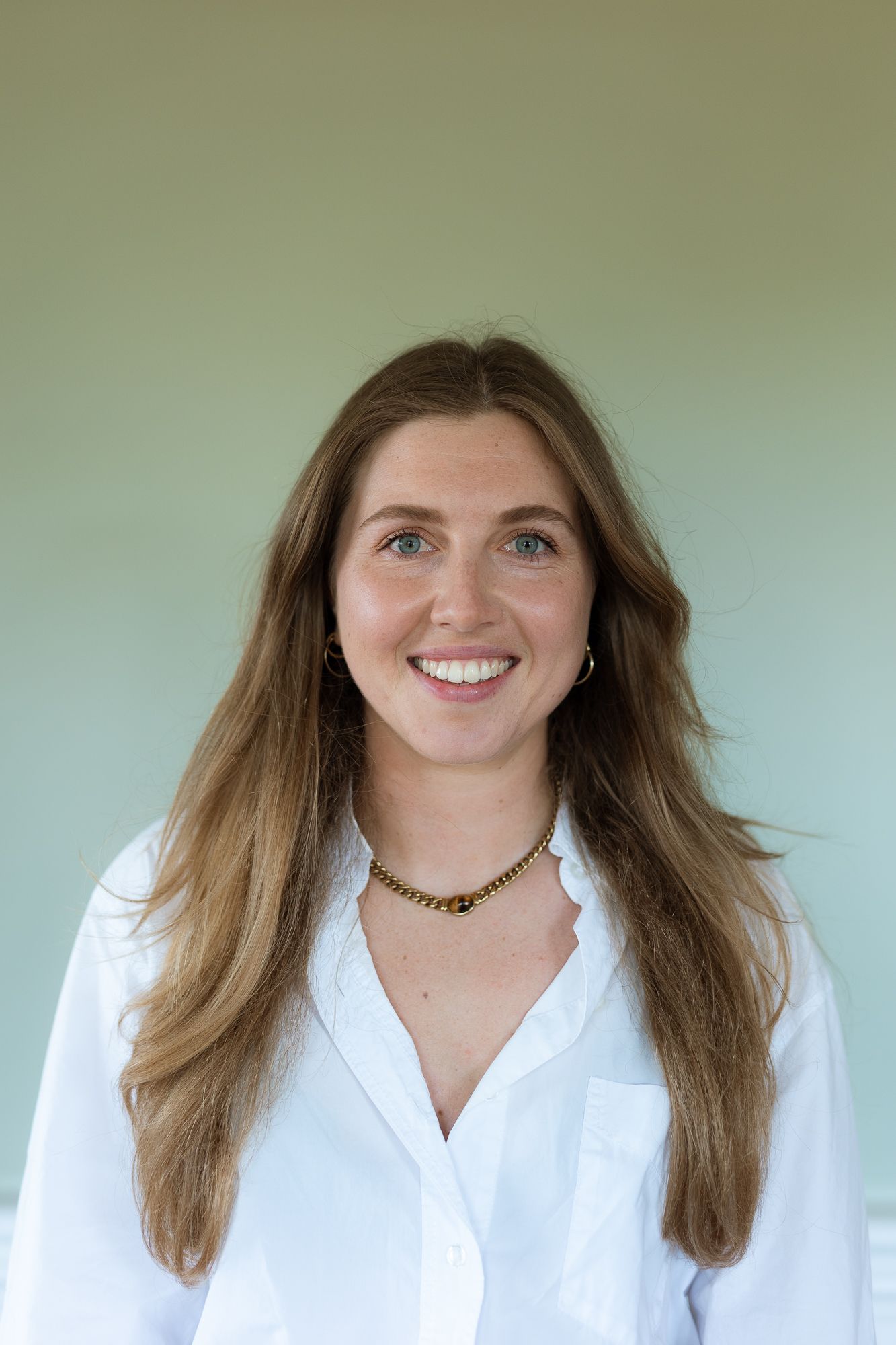 Smiling young woman with long brown hair wearing a white button-up shirt and gold necklace against a green background.