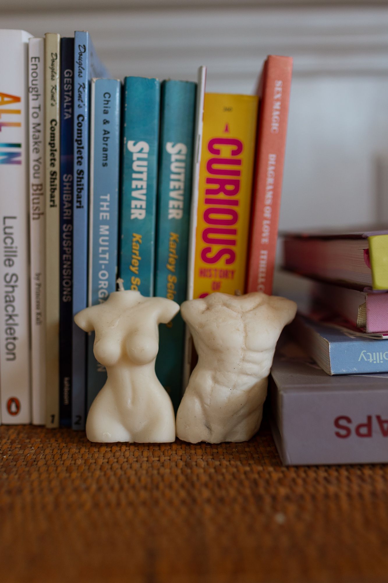 Two white body-shaped candles displayed as bookends on a shelf with colorful books including Curious.