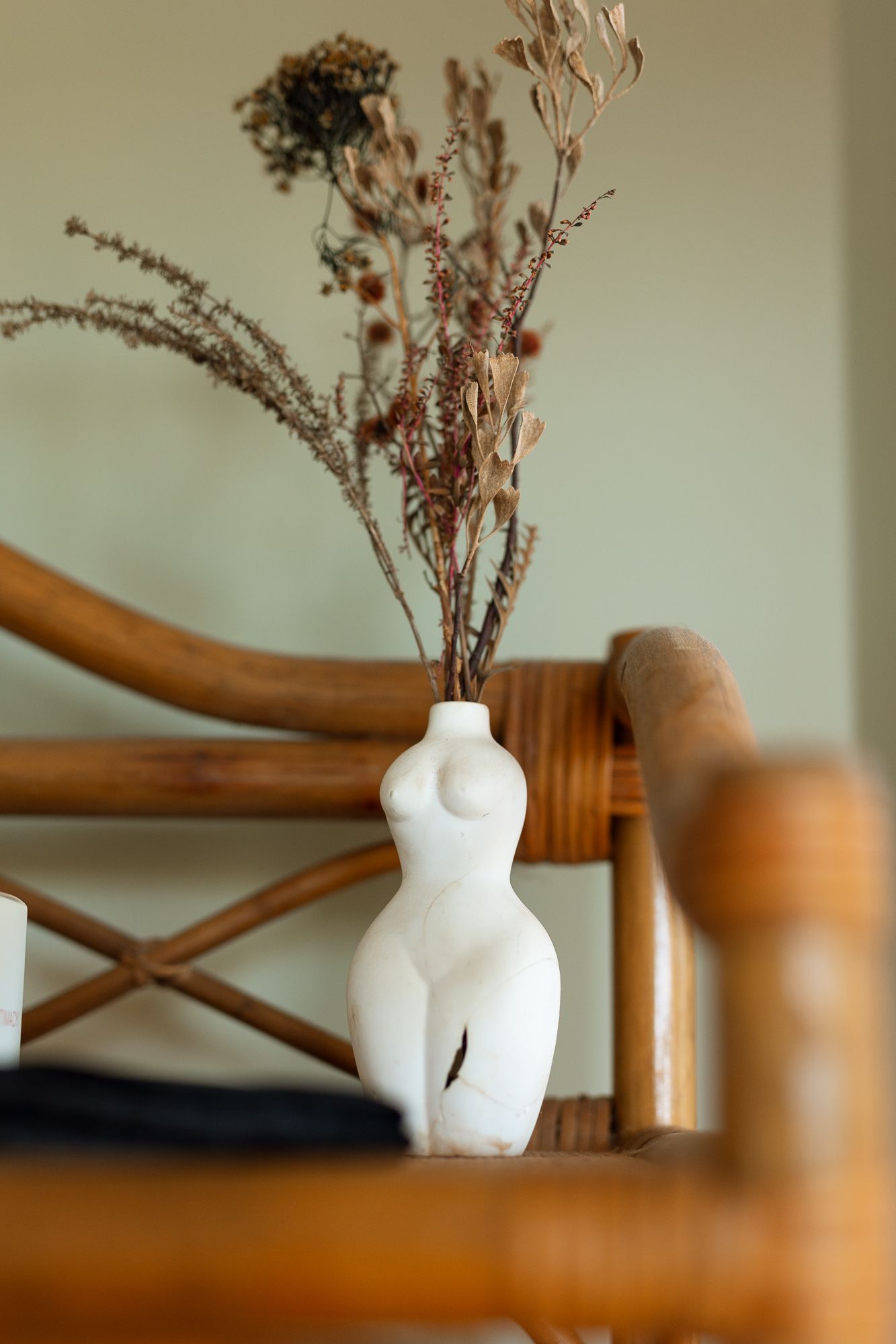 White ceramic female body vase with dried flowers on wooden rattan chair against sage green wall.