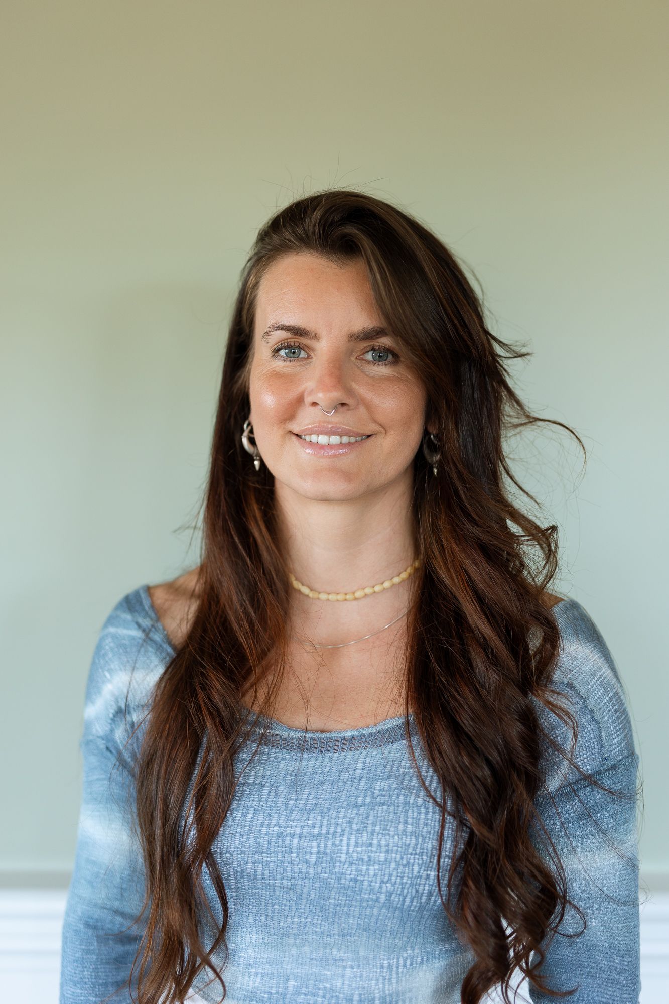 Smiling woman with long dark hair wearing a blue top and gold necklace standing against a light green wall.