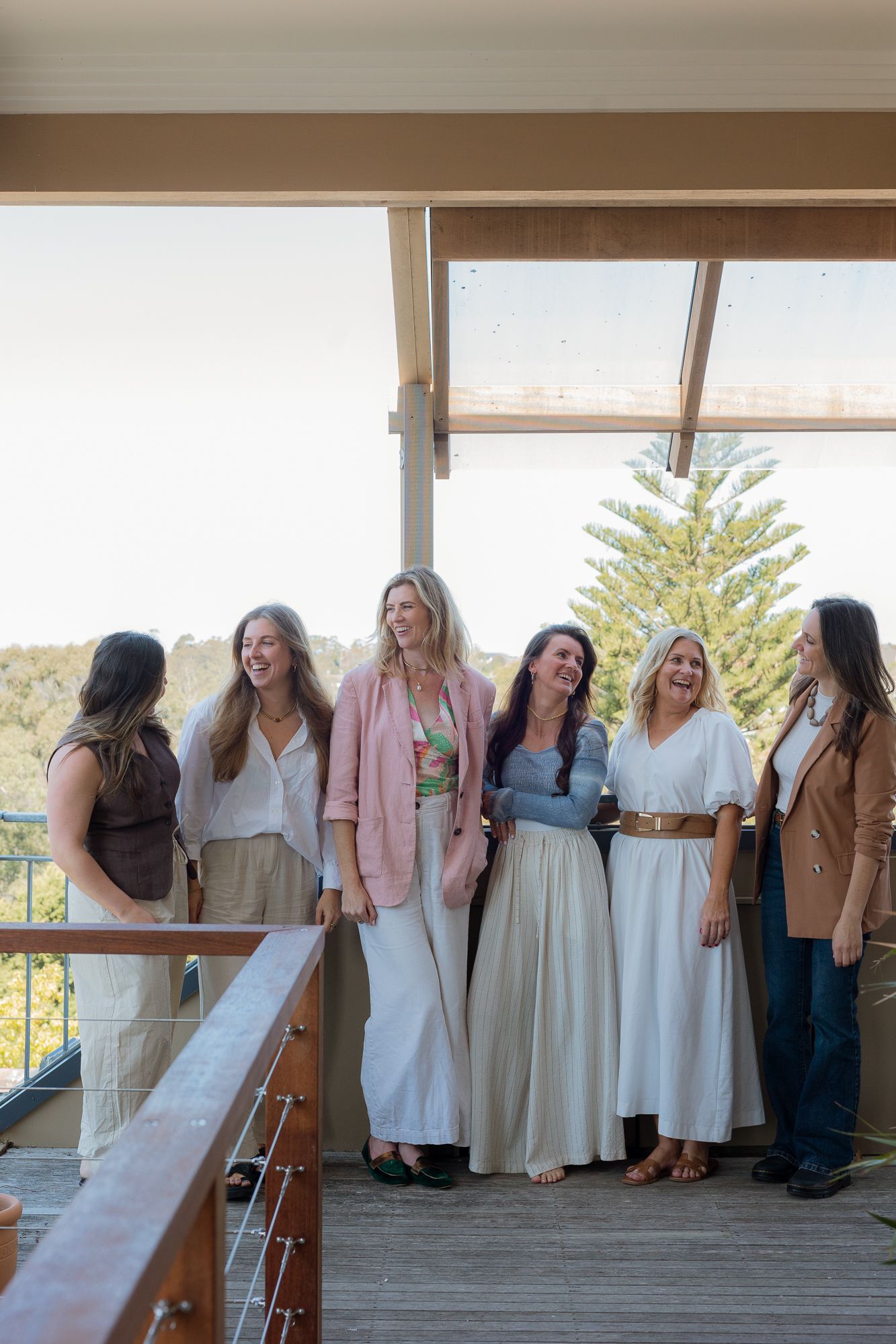 Six women laughing together on a wooden deck overlooking trees, dressed in elegant casual attire at an outdoor event.