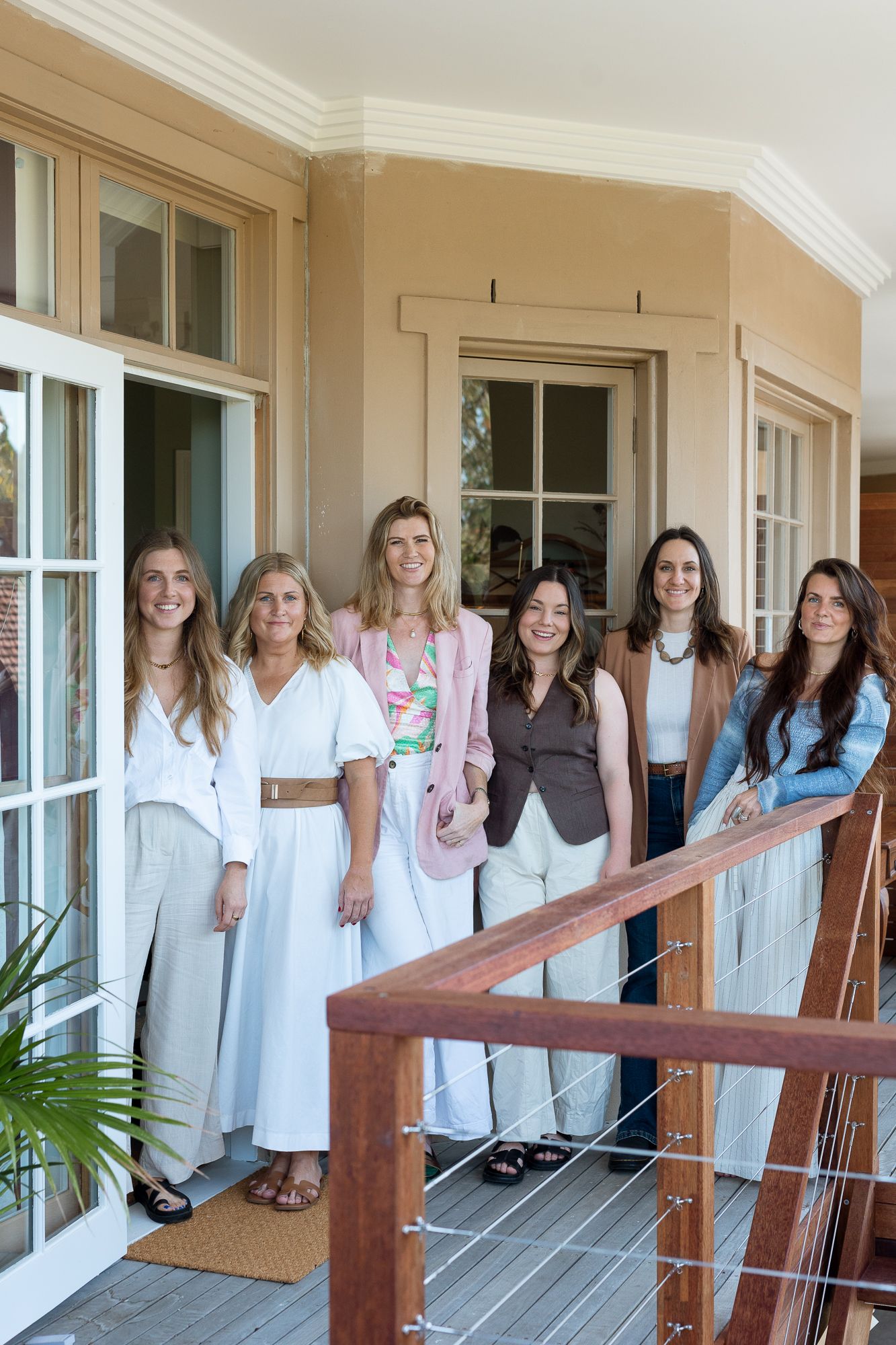 Six smiling women posing together on a wooden balcony outside a classic home.